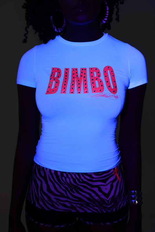 A model wearing a UV-reactive short-sleeve t-shirt that glows bright blue under blacklight, featuring the word 'BIMBO' in pink with diamantes across the chest, paired with zebra-print shorts.